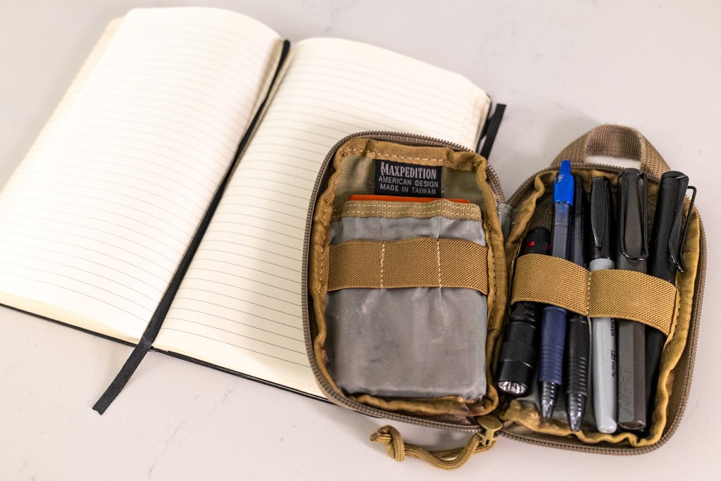 Maxpedition Micro Organizer open beside a Moleskine notebook with Lamy Safari pens, a Sharpie and folding readers